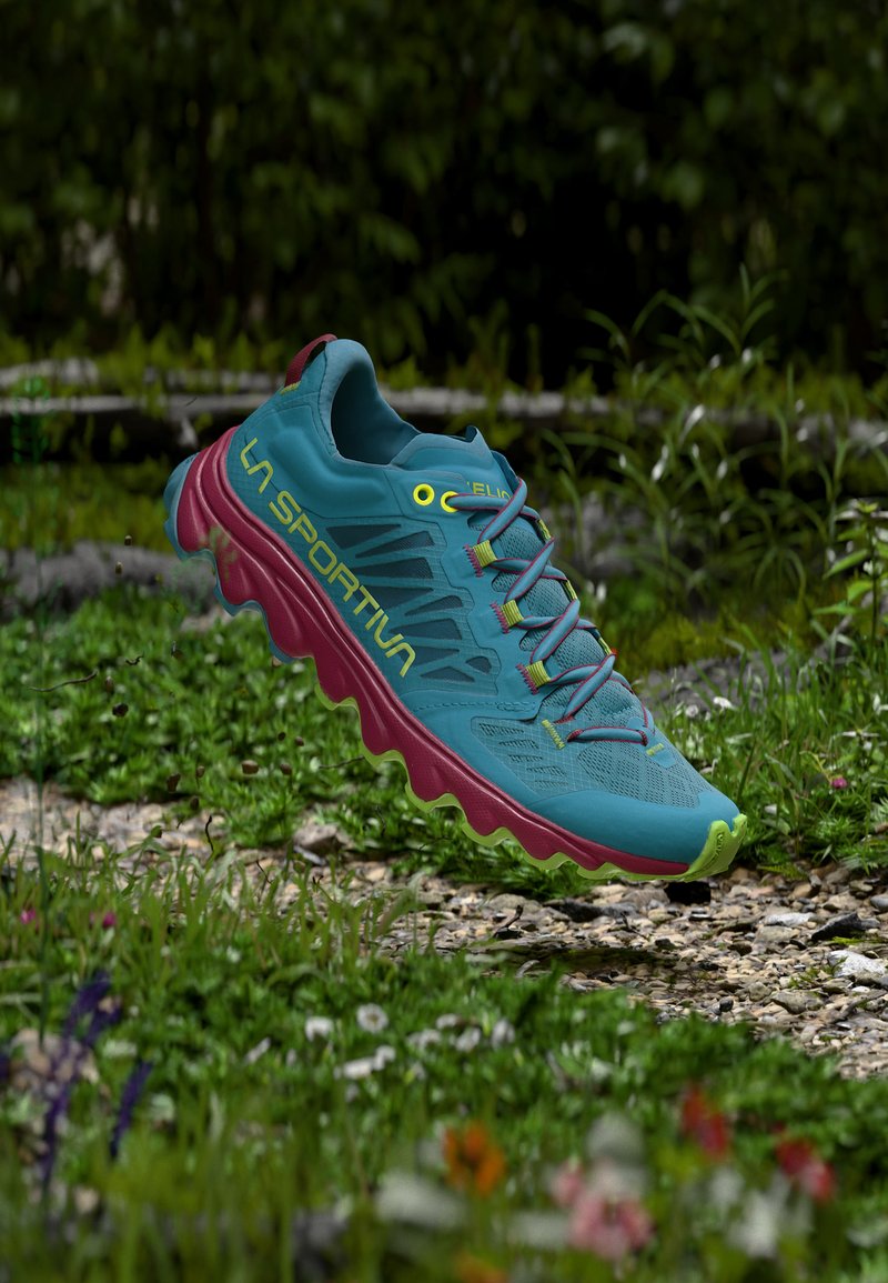 Chaussure de trail running bleue et rose avec des accents jaunes flottant au-dessus d'un sentier rocheux entouré d'herbe verte et de fleurs sauvages.