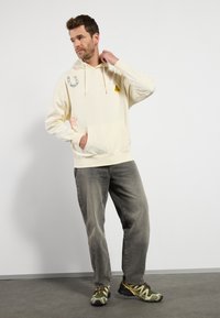 Creme hoodie med tegneserie-patches, frontlomme og snøre. Bæres med grå jeans og mønstrede sneakers, sat mod en ensfarvet baggrund.