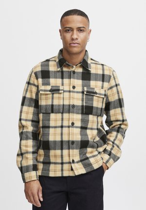 Flannel shirt in beige and black plaid, featuring two buttoned chest pockets, a collar, and dark buttons on a soft, textured fabric.