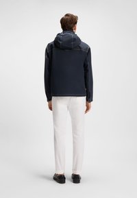 Tamsiai mėlynas striukės gobtuvas su kontrastingais baltais siūlais, dėvima ant baltų kelnių. Dizainas pasižymi laisvu siluetu ir raišteliu gobtuve.