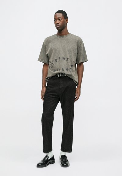 Tricou oversized gri cu text grafic, manșete uzate. Blugi negri cu talie înaltă, mocasini negri strălucitori și șosete albe. Design minimalist.