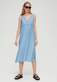 Jasnoniebieska sukienka dżinsowa z dekoltem w serek, bez rękawów, z bocznymi rozcięciami i regulowanymi sznurkami. Noszona z czarnymi sandałami.