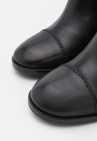 Gros plan sur des bottes en cuir noir lisse avec des orteils ronds et une couture visible sur fond blanc.