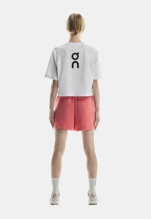 Personne debout de dos, portant un t-shirt court blanc avec un logo noir, un short corail, des chaussettes blanches et des baskets beiges.