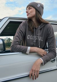 Sweat à capuche marron avec le texte "coastal" en blanc, manches longues, et bonnet en maille gris. Vu en appui contre un véhicule blanc avec des accents chromés.