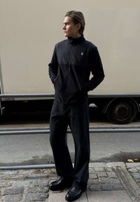 Mladý muž v čiernej bunde na zips, široké čierne nohavice a čierne topánky stojaci na dláždenej ulici vedľa béžového nákladného auta.