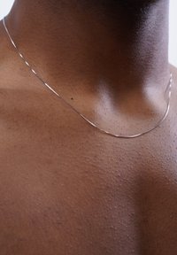 Dünne Silberkette, die auf dem Schlüsselbein liegt, mit glatter Textur und dezentem Glanz auf brauner Haut.