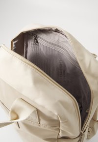 Sac à dos en tissu beige avec fermeture zippée. L'intérieur comprend une poche en mesh et un revêtement lisse. Design simple, sans motifs ni accents visibles.