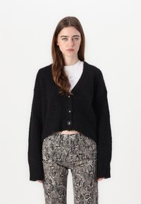 Cardigan nero, corto, con un motivo in maglia testurizzata e quattro grandi bottoni. Abbinato a pantaloni aderenti con una fantasia in stampa serpente.