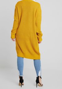 Cárdigan largo y holgado en un amarillo brillante, de punto texturizado. Cuenta con puños y dobladillo de canalé, combinado con jeans desgastados azul claro y tacones negros.