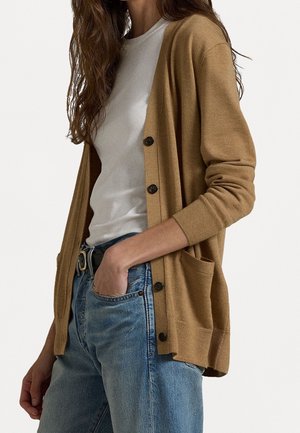 Vrouw die een wit T-shirt draagt, een beige vest met knopen en zakken, en een blauwe spijkerbroek met een zwarte riem, hand in de zak, tegen een effen achtergrond.