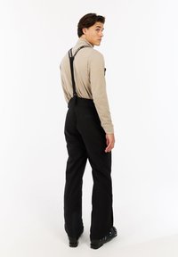 Zwarte skibroeken met verstelbare zwarte bretels, een gladde textuur en ritszakken. Gdragen over een lichtbeige lange mouw top.