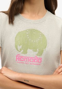 Lys grå t-shirt med et grønt elefantgrafik med indviklede mønstre og pink tekst nedenfor, der står "Komodo London Bali Kathmandu."