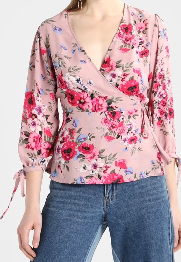Blouse cache-cœur fleurie rose avec manches trois-quarts bouffantes nouées aux poignets, associée à un jean en denim bleu.