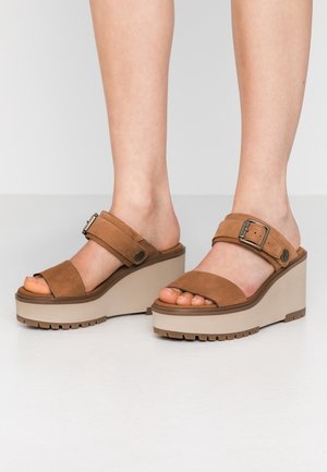 Pieds portant des sandales compensées en daim marron avec des brides réglables à boucle, sur un sol clair et un fond uni.