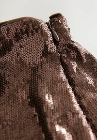 Detailní záběr hnědé látky se flitry s hnědým jezdcem zipu na oděvu na světlém pozadí.