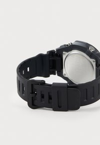 Montre en caoutchouc noir avec un bracelet texturé, un fond en métal rond et un boîtier épais. Elle possède un bouton sur le côté et une boucle robuste.