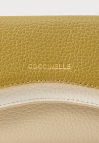 Artimųjų planas, kur matyti tekstūruota oda su įspaustu auksiniu "Coccinelle" logotipu ir smėlio bei baltos odos siūlėmis.