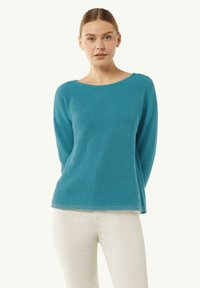 Jasnoniebieski sweter robiony na drutach z szerokim dekoltem, prążkowaną fakturą i długimi rękawami, w zestawieniu z białymi spodniami. Miękki, casualowy design.