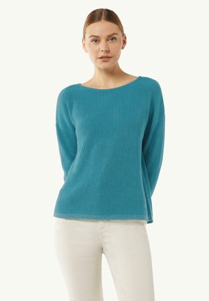 Jasnoniebieski sweter robiony na drutach z szerokim dekoltem, prążkowaną fakturą i długimi rękawami, w zestawieniu z białymi spodniami. Miękki, casualowy design.