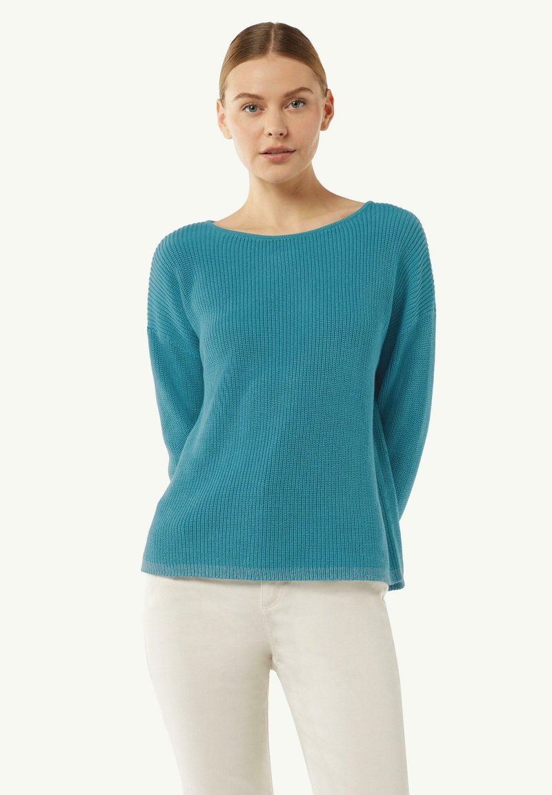 Jasnoniebieski sweter robiony na drutach z szerokim dekoltem, prążkowaną fakturą i długimi rękawami, w zestawieniu z białymi spodniami. Miękki, casualowy design.