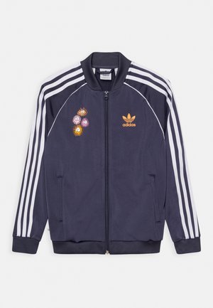 Chaqueta de chándal Adidas azul marino con rayas blancas en las mangas, cremallera frontal, logo de Adidas en naranja y parches de flores de dibujos animados coloridos en el lado izquierdo del pecho.