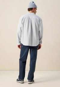 Man in een lichtblauwe gebreide beanie, een lichtgestreept overhemd met knopen, marineblauwe broek en donkere sneakers, staand met de rug naar de camera tegen een beige muur.