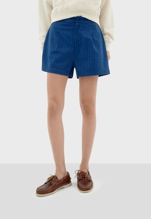 Navyblauwe katoenen shorts met een gemêleerd patroon, voorzien van een knoop aan de voorkant, zijzakken en een relaxed fit, gecombineerd met bruine boot schoenen.