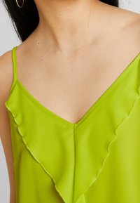 Top de satén verde con escote en V y detalle de borde con bordes ondulados, con tirantes finos y textura suave. Vista de cerca del escote y la tela.