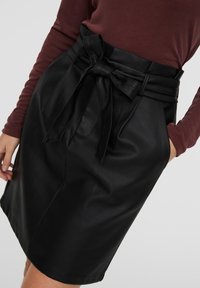 Falda de piel negra con cintura alta y lazo decorativo, bolsillos laterales y textura suave. Combinada con una blusa burdeos visible.