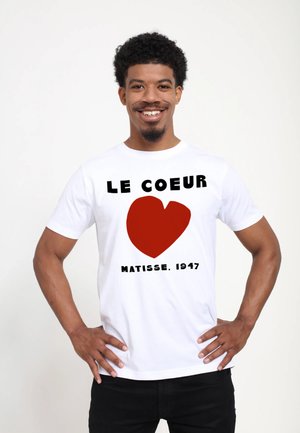 Homme souriant portant un T-shirt blanc avec un cœur rouge et le texte "Le Coeur Matisse, 1947", debout les mains sur les hanches.