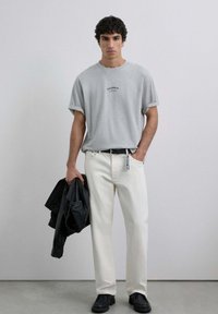 Camiseta gris de manga corta con puños enrollados, pantalones vaqueros blancos de pierna recta, chaqueta de cuero negra en la mano y zapatos negros. Diseño simple y limpio.