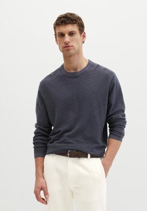 Młody mężczyzna z krótkimi włosami, ubrany w ciemnoszary sweter z długim rękawem, białe spodnie i brązowy pasek, stojący na jednolitym tle.