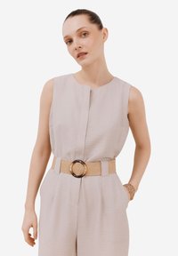 Ljusbeige ärmlös jumpsuit med rak skärning, som har ett texturerat tyg och ett vävt brunt bälte med en rund spänne.