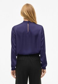 Mørk lilla bluse med lange ermer, rynket halslinning og nøkkelhull-detalj bak, kombinert med svarte bukser med elastisk midje. Jevn stofftekstur.