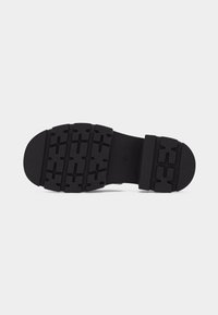 Semelle de chaussure noire avec un motif de bande de roulement rectangulaire texturé et un talon épais, présentée sur un fond gris clair uni.