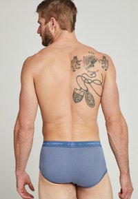 Blå bomullsboxer med en snug passform och en Jockey-logotyp på midjan. Bakifrån syns en tatueringdesign på den övre delen av ryggen.