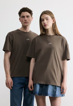 Fiatal férfi és nő oldalról állnak túlságosan nagy barna pólókban; a nő kék farmerszoknyát, a férfi kék farmert visel.