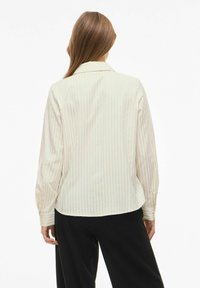 Vrouw met lang bruin haar draagt een crèmekleurige blouse met streepjes en zwarte broek, met de rug naar de camera gericht tegen een eenvoudige witte achtergrond.