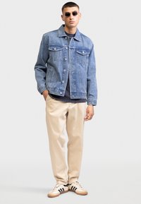 Man met zonnebril, een blauwe spijkerjack, donkerblauw shirt, beige broek en witte sneakers met zwarte strepen, staand tegen een effen achtergrond.