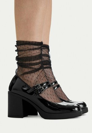 Piernas con calcetines tobilleros negros transparentes con lunares y bordes con volantes, combinados con zapatos de charol negro brillante estilo Mary Jane con doble correa.