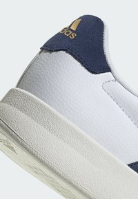 Bijele kožne tenisice s mornarsko plavim suede petnim dijelom i zlatnim logotipom. Teksturirana gumena potplat s laganim uzorkom za bolji grip.