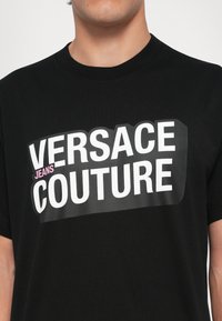 Svart T-skjorte i bomull med en dristig, trykt hvit tekstlogo "VERSACE JEANS COUTURE" med en rosa aksent på "JEANS." Rund halslinning.