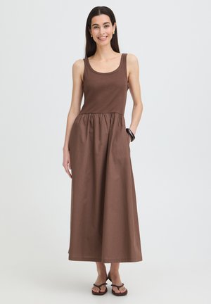 Mujer sonriente, vestida con un vestido maxi marrón sin mangas con cintura fruncida, pulsera negra, pendientes dorados y sandalias negras sobre un fondo liso.