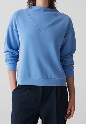 Personne portant un pull bleu clair à manches longues avec une couture en forme de V et un pantalon noir, debout avec une main dans la poche devant un fond uni.