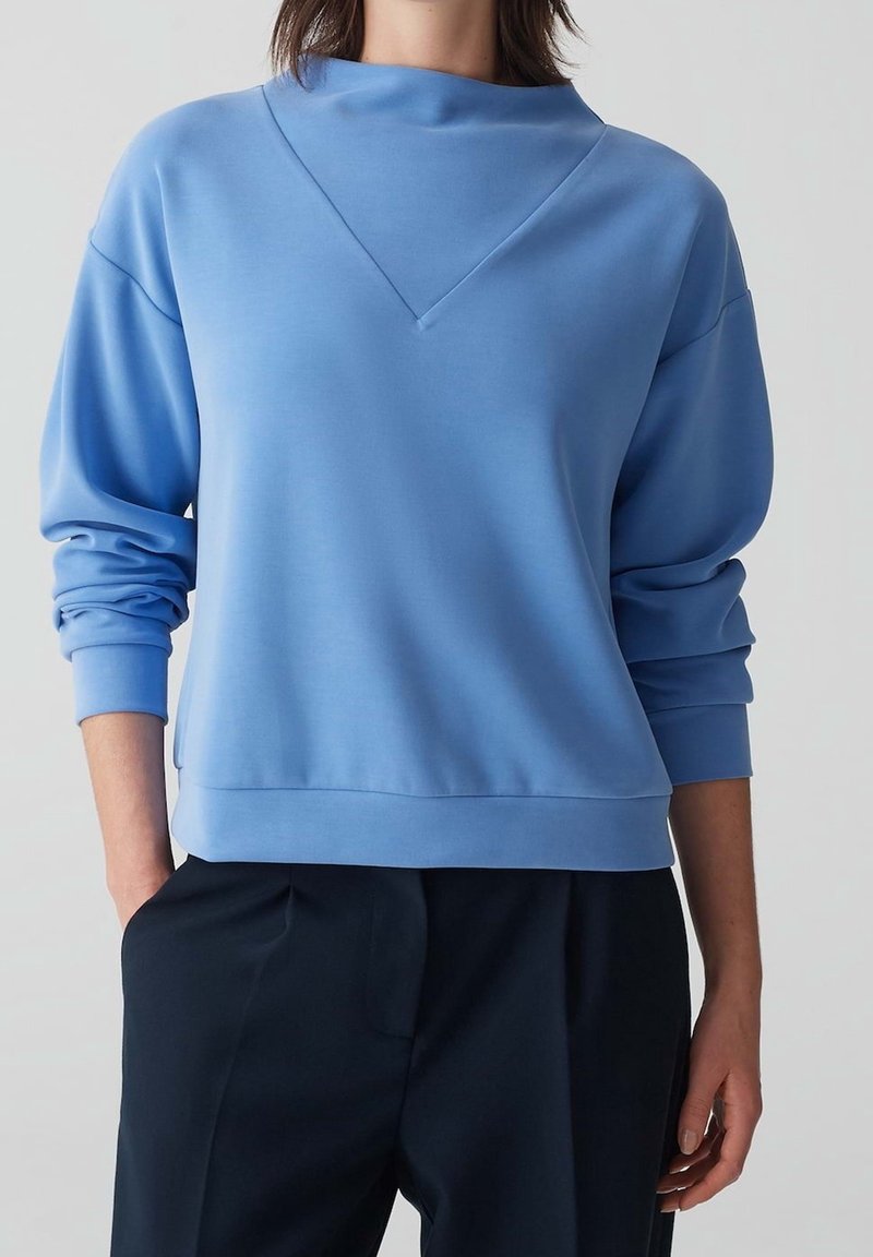 Personne portant un pull bleu clair à manches longues avec une couture en forme de V et un pantalon noir, debout avec une main dans la poche devant un fond uni.