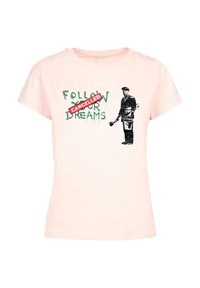 Lyserød T-shirt med korte ærmer, med sorte grafikker af en figur, der holder en pensel, og tekst der siger "FØLG DINE DRØMME" med "AFLYST" i rødt.