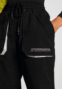 Svarta cargobyxor med elastisk midja, reflekterande detaljer och vit broderad logotyp på fickan. Materialet ser slätt och hållbart ut.