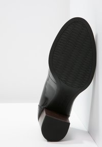 Bottine en cuir noir avec un talon rectangulaire en bois, une semelle en caoutchouc noir texturée et une finition lisse sur la tige avec des coutures minimales.