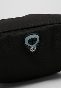 Pochette en tissu noir à surface texturée ; présente un logo bleu et noir sur le côté, une forme triangulaire et une fermeture éclair.